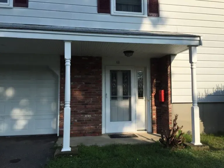 A residential entrance with a glass door framed by red brick and white siding. Two white pillars support the porch roof. A garage is visible to the left.