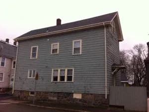 Two-story gray house with multiple windows, stone foundation, and peaked roof on a cloudy day. Sparse trees are visible in the background.