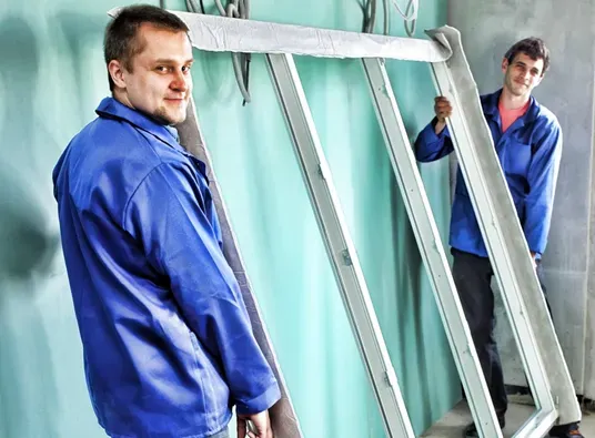 Two men in blue work jackets smiling while carrying a large window frame indoors. The atmosphere is light and cooperative, suggesting teamwork.