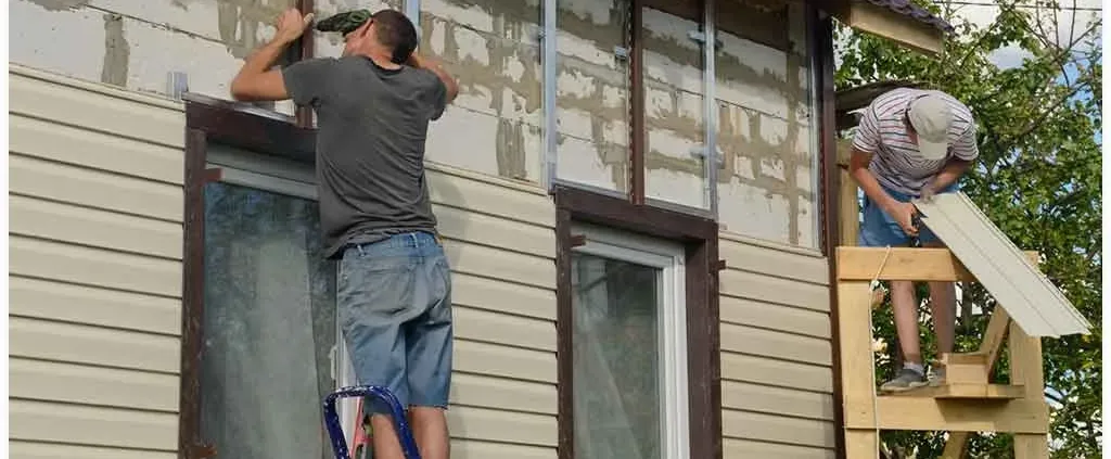 Two men are installing beige siding on a house. One is on a ladder and the other on a scaffold. The scene conveys teamwork and home renovation.