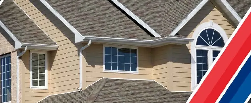 A beige house with a steeply pitched roof and multiple windows. A red, white, and blue diagonal stripe design is on the bottom right corner.