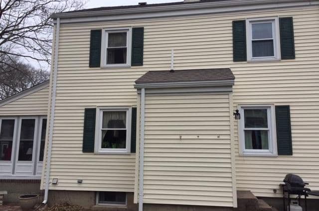 A beige house with white trim and dark green shutters. Central small porch, windows on the first and second floors. Overcast sky, bare tree branches.