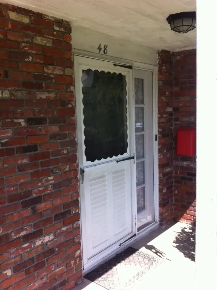 Entrance with a white metal screen door on a red brick wall, numbered 48. A red mailbox is nearby, and a black outdoor light fixture hangs above.