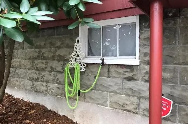 A green garden hose hangs neatly on an ornate white hook against a stone-textured wall beneath a small window. Nearby, a red gutter descends beside foliage.