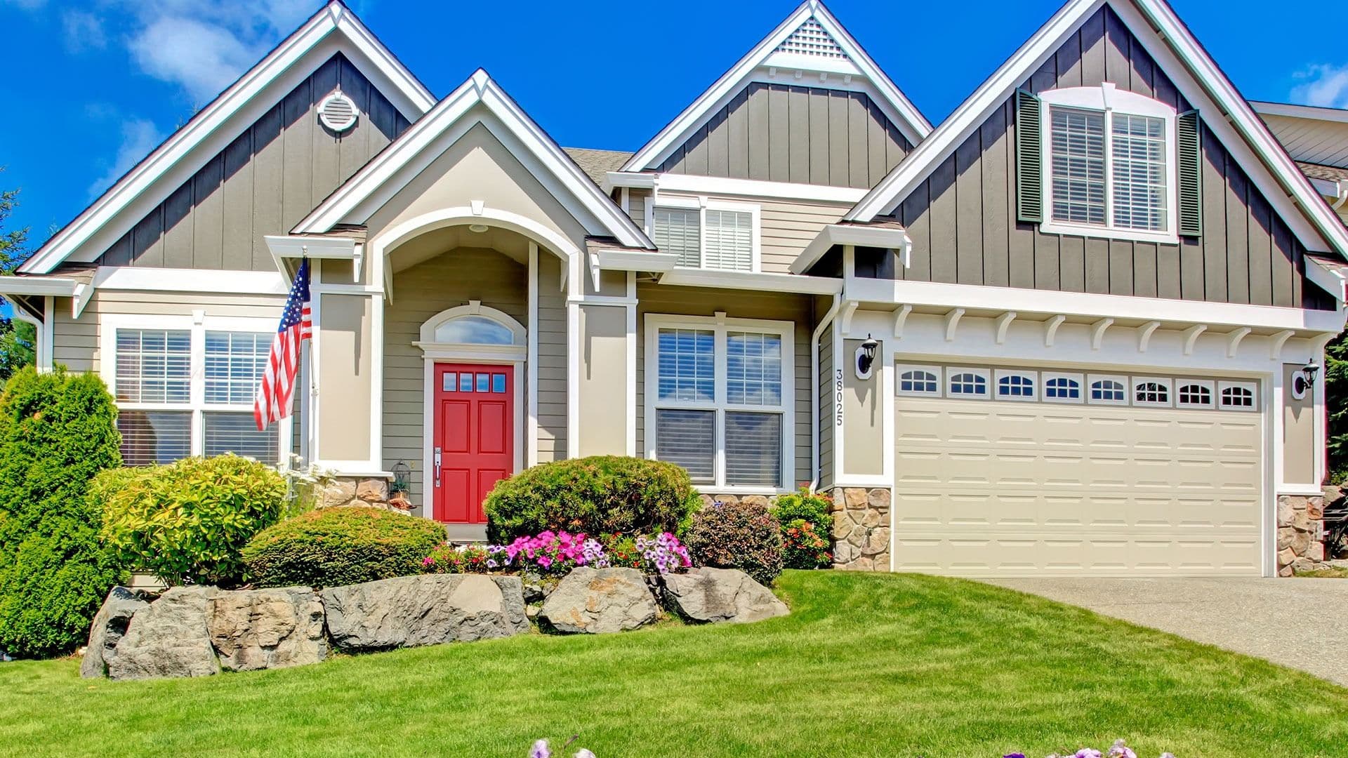 Pale yellow house with red door and American flag with well landscaped yard.