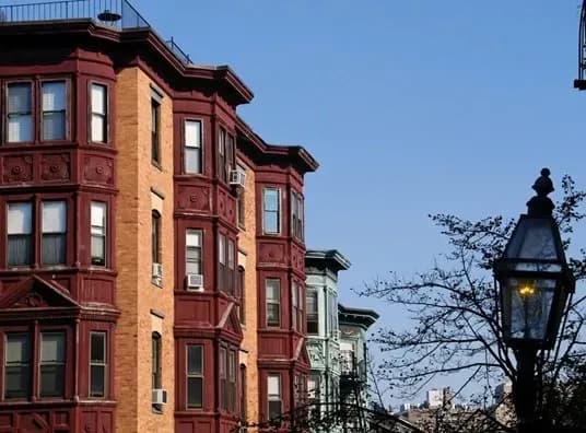Historic brownstone buildings with detailed facades in a city neighborhood stand against a clear blue sky. A vintage streetlamp glows softly nearby.