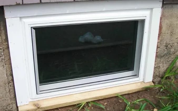A basement window with a white frame set into a concrete wall. The glass reflects a dark interior. Green plants surround the base, adding a natural touch.