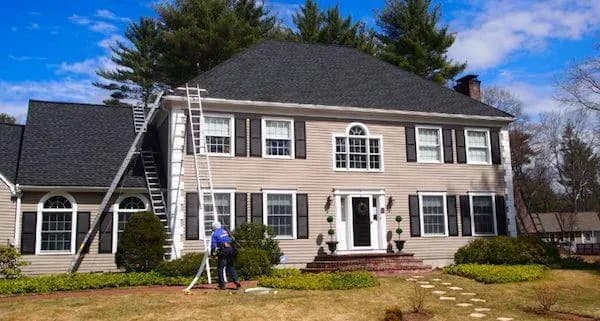 Two-story colonial house with beige siding, black shutters, and a dark roof. A person on a ladder works on the facade amid a neat front yard. Clear sky above.