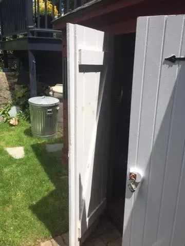 A gray wooden shed door stands partially open in a sunlit garden. Nearby, a metal trash can is on green grass, with a stone path leading to a dark step.
