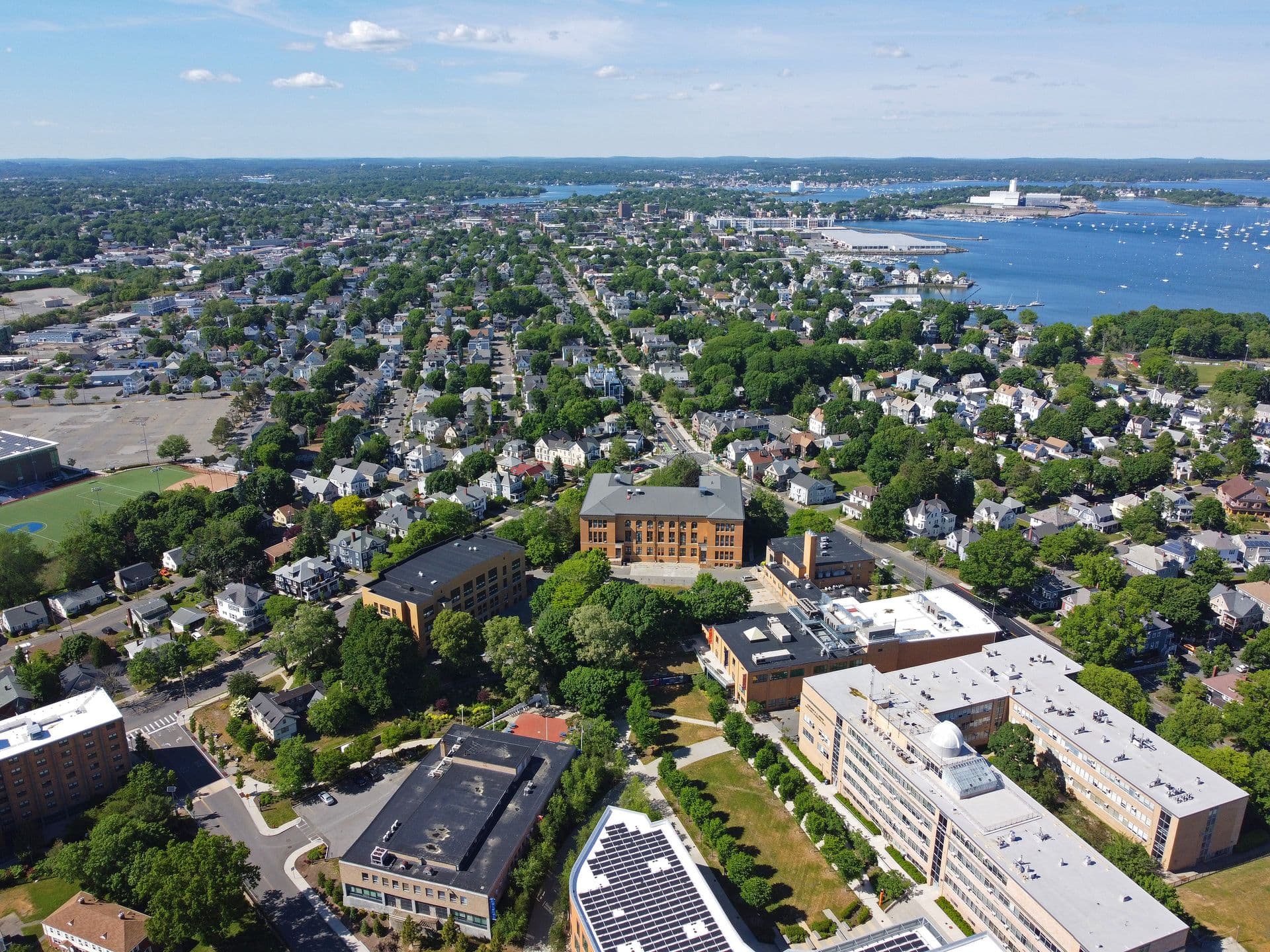 Aerial view of a coastal city with dense greenery and buildings. In the distance, there are docks and boats on calm blue waters under a bright sky.