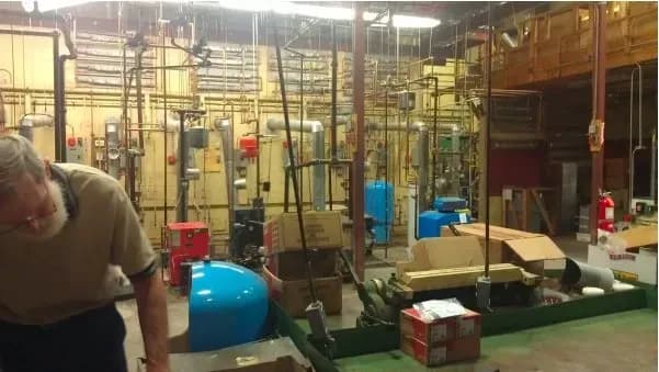 A cluttered industrial room with pipes, machinery, and boxes. A person bends over on the left, suggesting maintenance or repair work is underway.