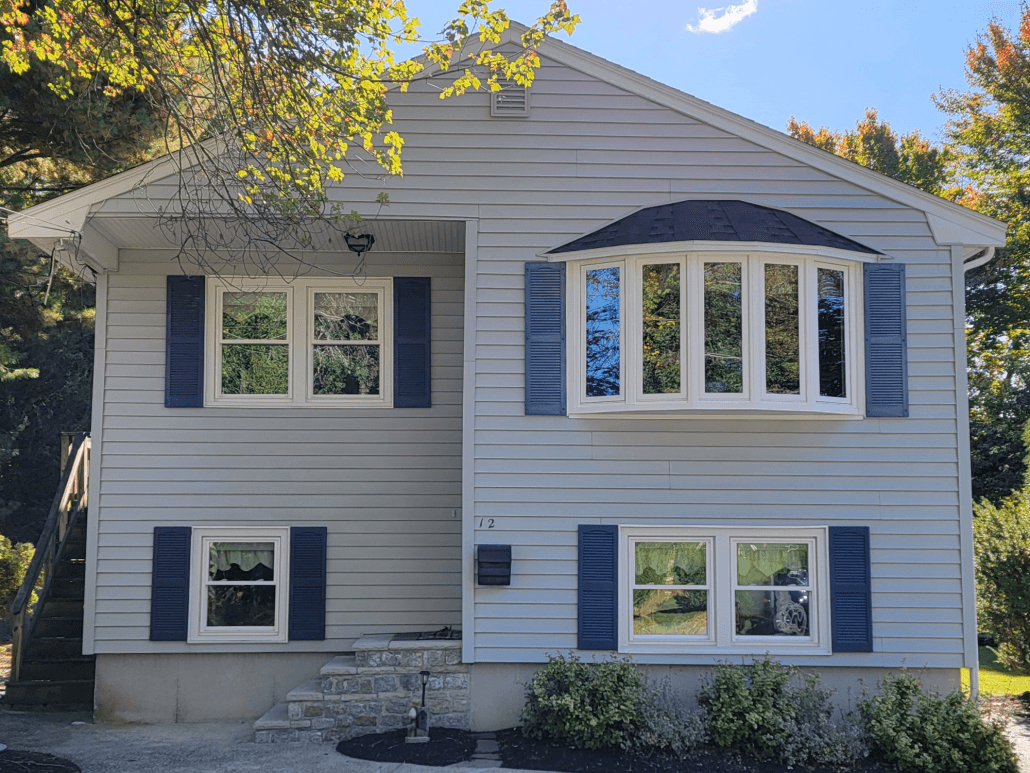 Two-story house with light gray siding and dark blue shutters. Features a prominent bay window on the right side. Greenery and shadows convey a serene atmosphere.