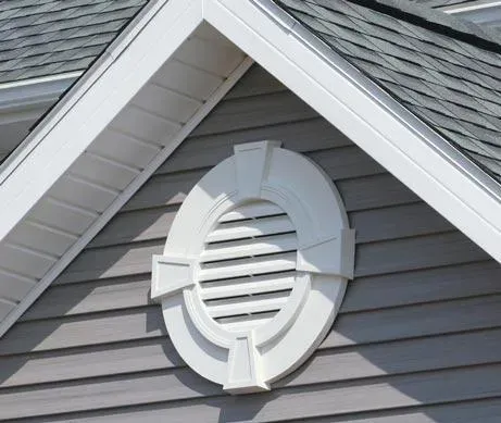 A white decorative vent with a circular design on the gable of a house, featuring gray siding and shingle roofing, under a clear sky.
