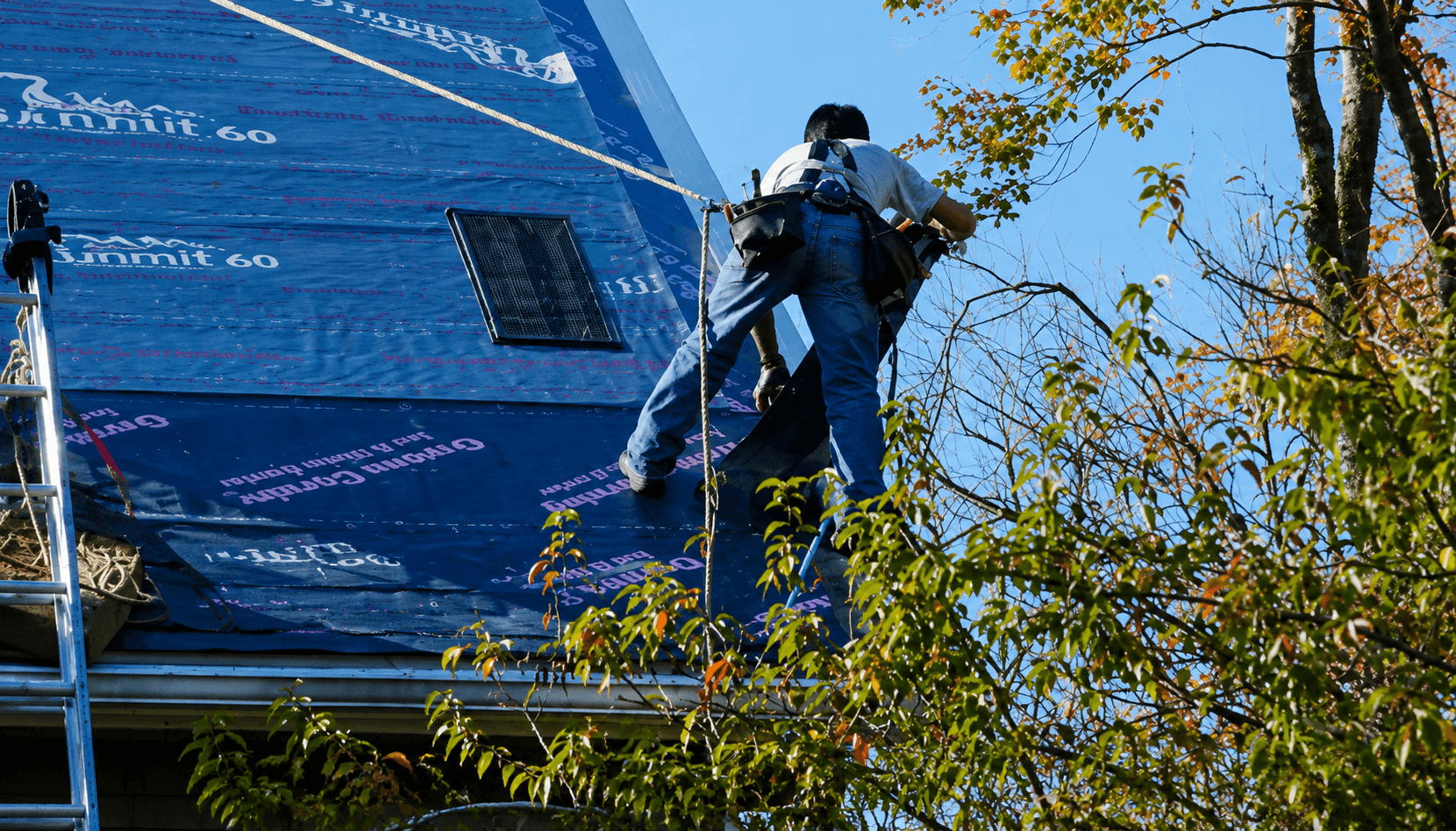 A person in harness repairs a steep roof covered in underlayment. A ladder is visible, with surrounding autumn trees, under a clear blue sky.