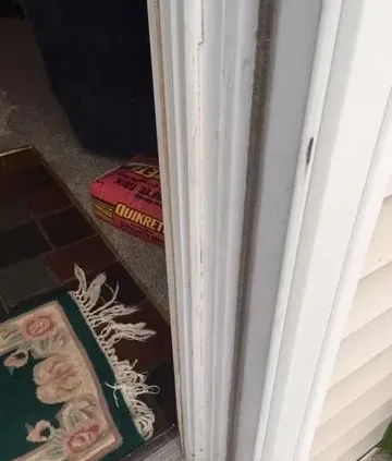 A bag of Quikrete lies wedged under a door slightly ajar, blocking it. Nearby, a fringed floral rug is on a tiled floor, suggesting a home setting.