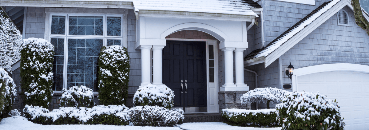 Snow-covered suburban house with a black front door, large windows, and bushes, conveying a peaceful, wintery atmosphere.