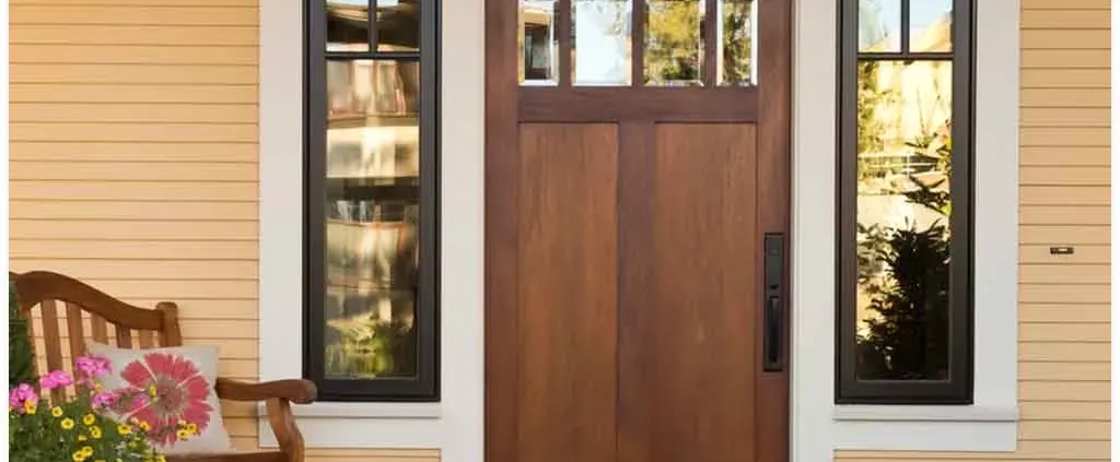 A wooden front door flanked by two tall windows is set in a yellow paneled wall. A bench with pink flowers adds a welcoming, homely touch.