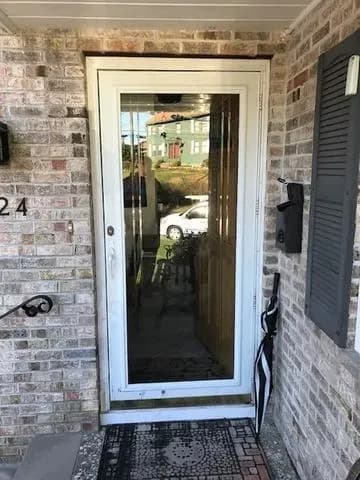 Front door with white frame and glass panel reflecting a car and house. The surrounding brick wall adds a cozy, welcoming feel. An umbrella leans nearby.