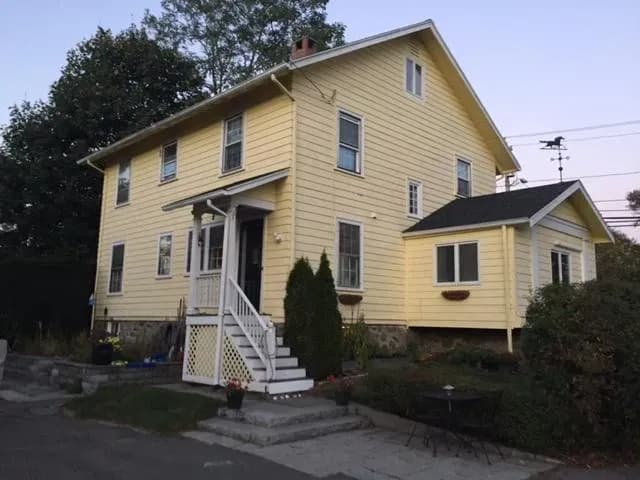 A charming two-story yellow house with a small porch and white railing at dusk. The yard features shrubs and a patio set, conveying a cozy, inviting feel.
