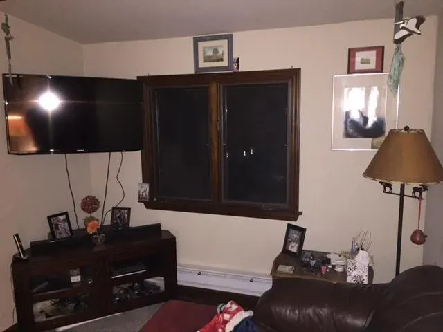 A cozy living room with a mounted TV, framed pictures, and a window at the center. A table lamp and decorations create a warm, inviting atmosphere.