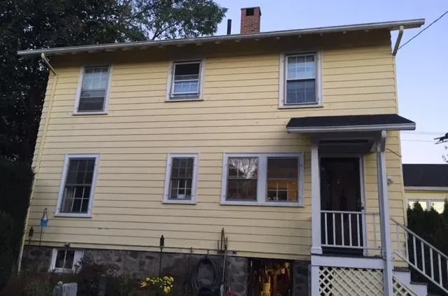 A two-story yellow house with white trim, featuring a small covered porch and six windows, sits on a stone foundation. The mood is calm and residential.
