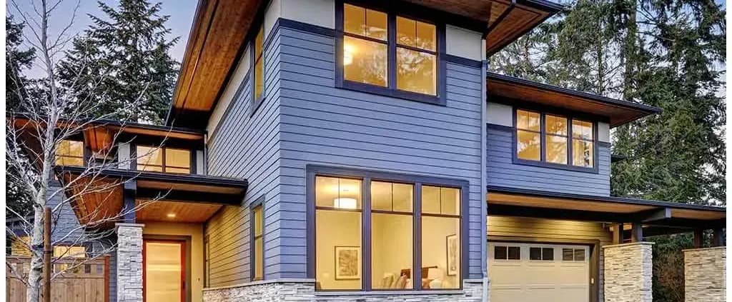 Modern two-story house with blue wood siding, large windows, and stone accents. Warm interior lighting visible, surrounded by tall trees. Serene atmosphere.