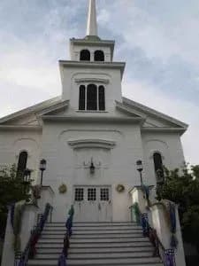 White church with a tall steeple against a cloudy sky. The front steps are decorated with colorful fabric, creating a welcoming and serene atmosphere.