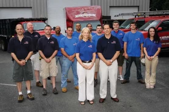 A group of eleven people in matching blue shirts stands outdoors in front of red company trucks and a building with white garage doors, smiling confidently.