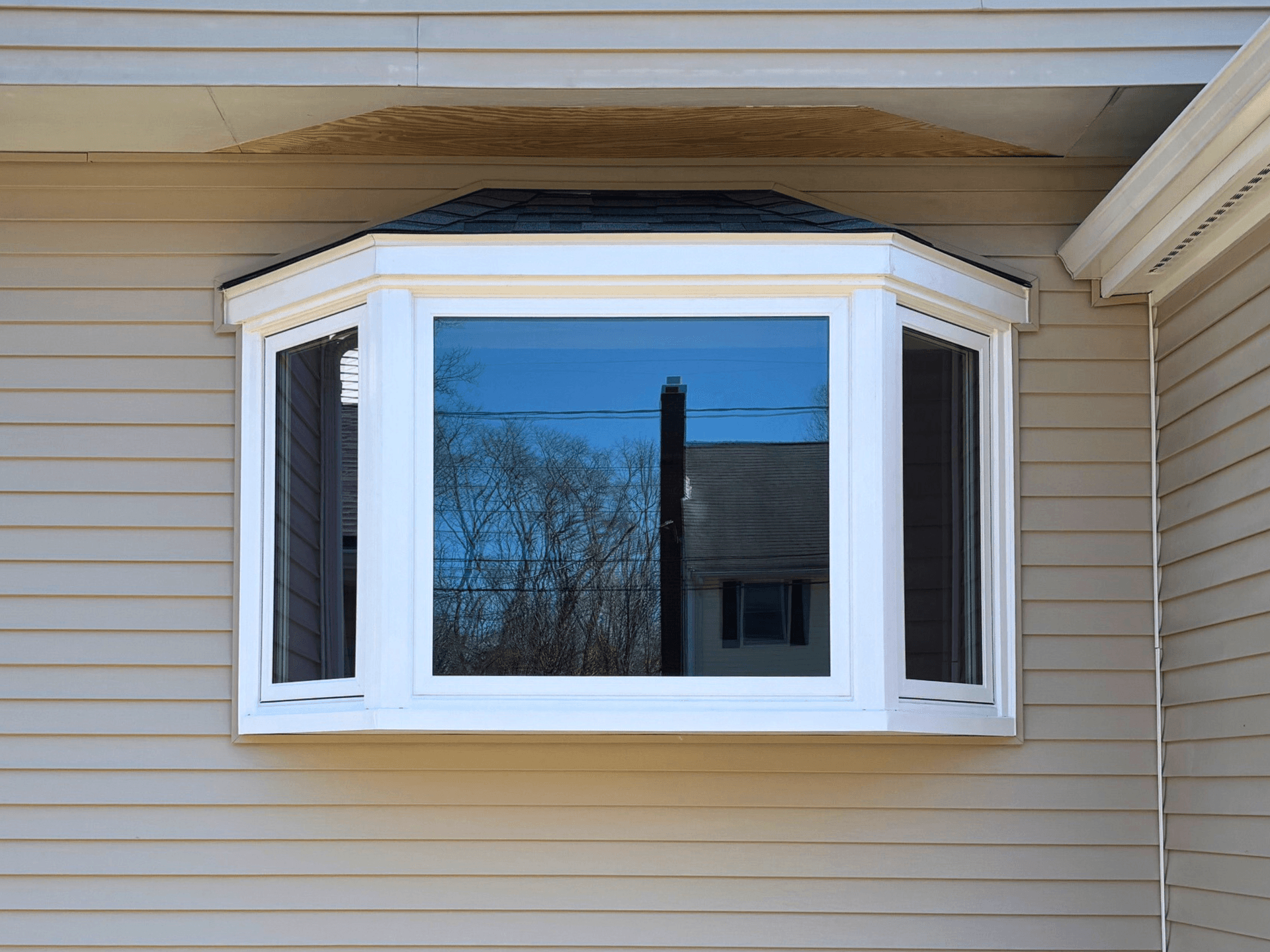 Bay window on a house with beige siding reflects trees and power lines on a clear day. The window has three glass panels framed in white.
