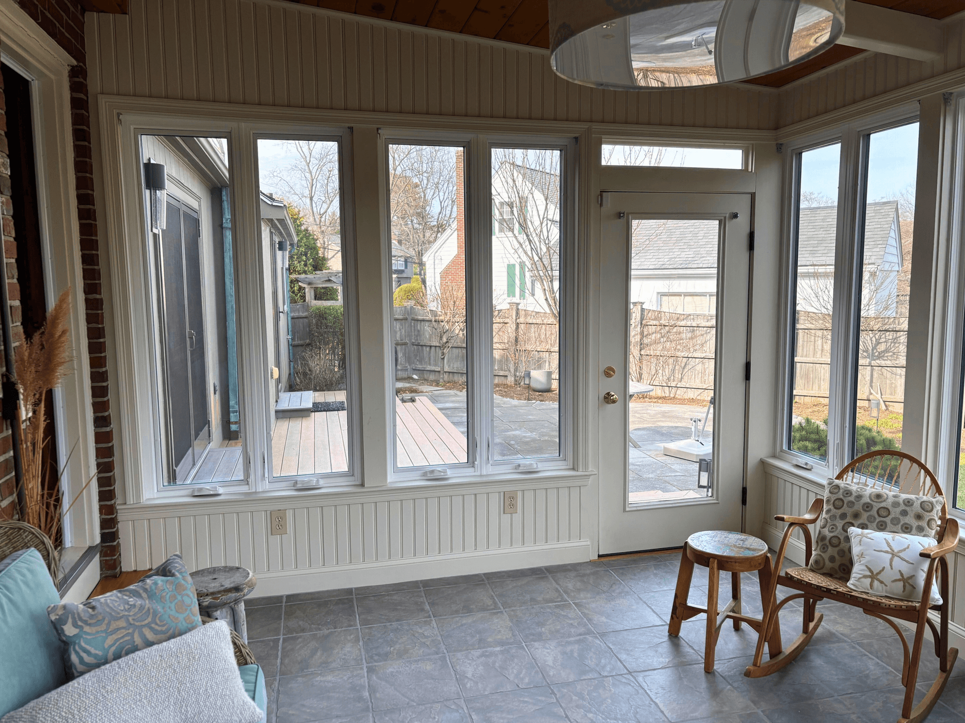 Sunlit porch with large windows and a glass door overlooking a backyard. Wooden chair with a starfish pillow and stool adds a cozy touch.