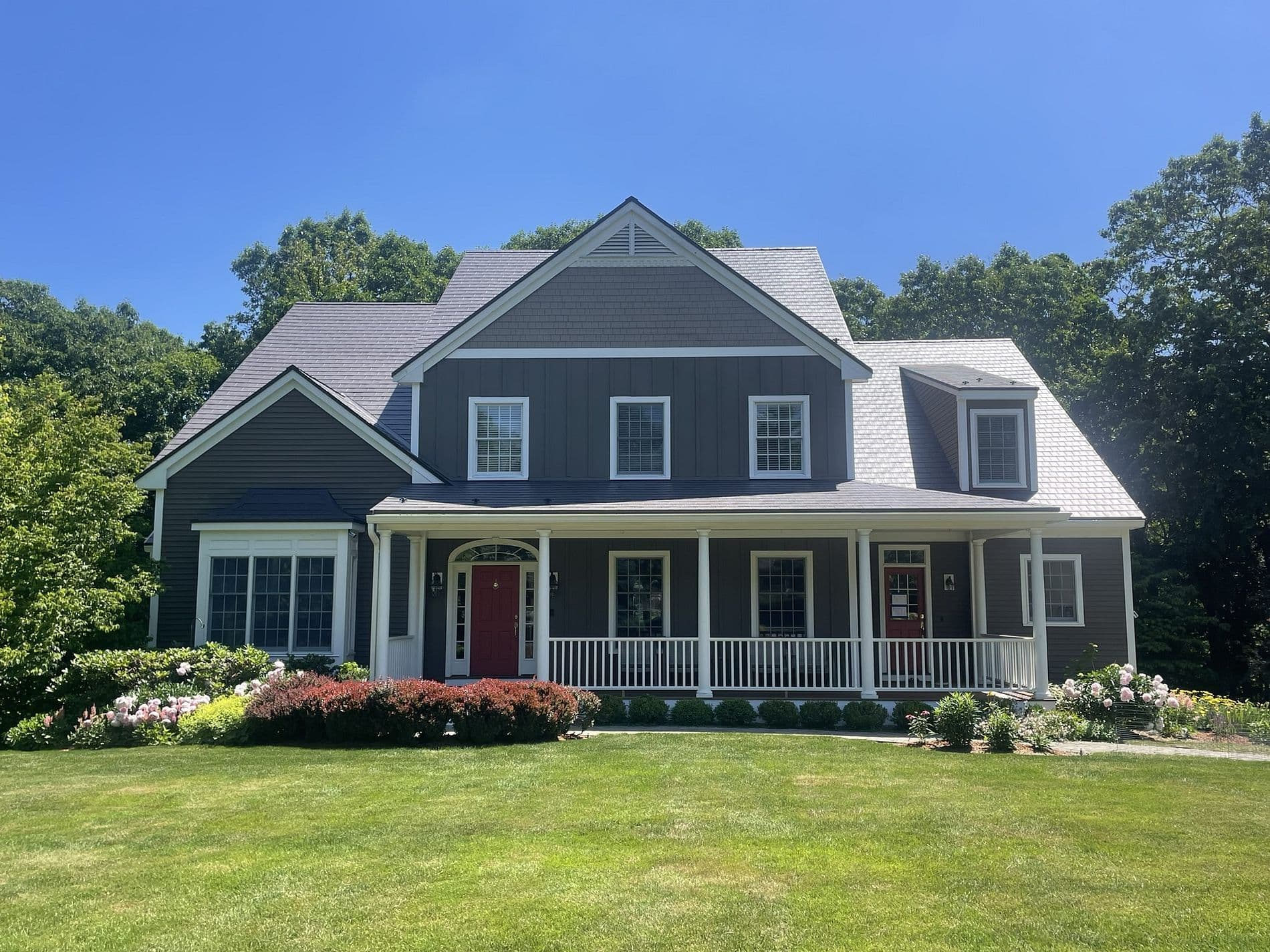 A charming gray house with a steep gabled roof and white trim sits in a lush green yard. It features a welcoming porch, red doors, and vibrant flowerbeds.