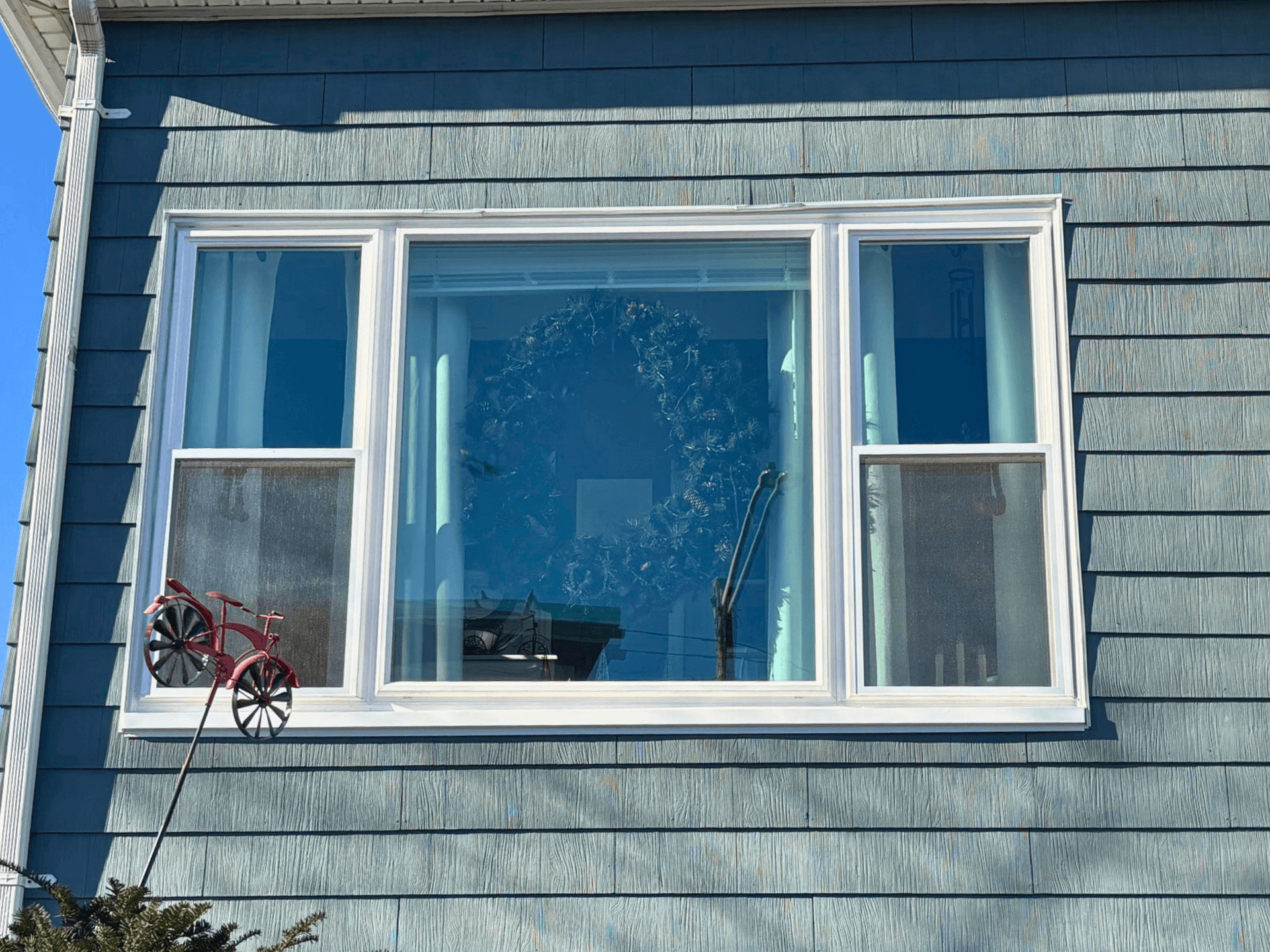 A blue house wall with a large window featuring a decorative wreath inside. Outside, a small red metal tricycle sculpture is mounted near the window.