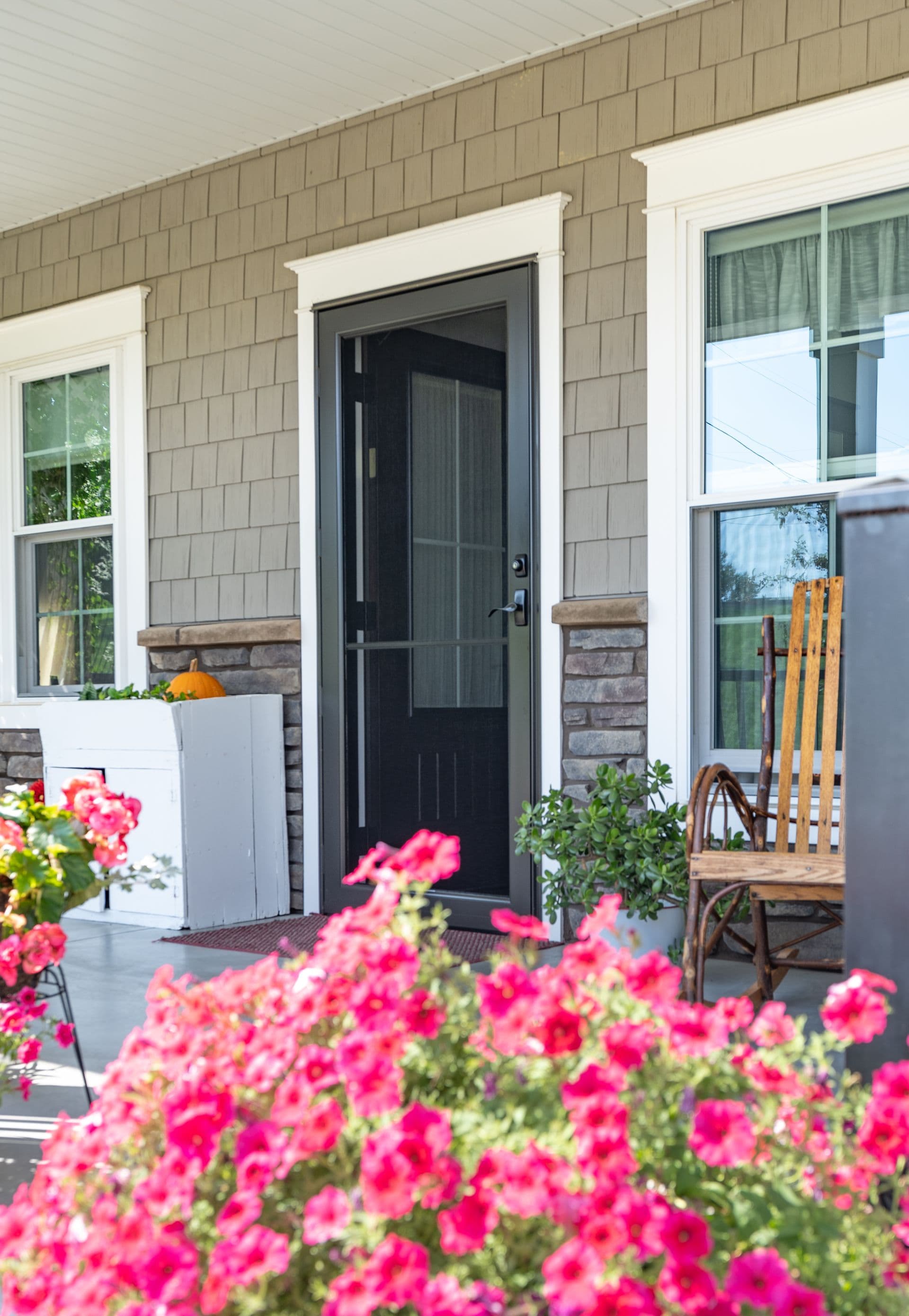 Cozy porch with a black door, wooden rocking chair, and vibrant pink flowers. A small pumpkin sits on a white box, creating a welcoming autumn feel.