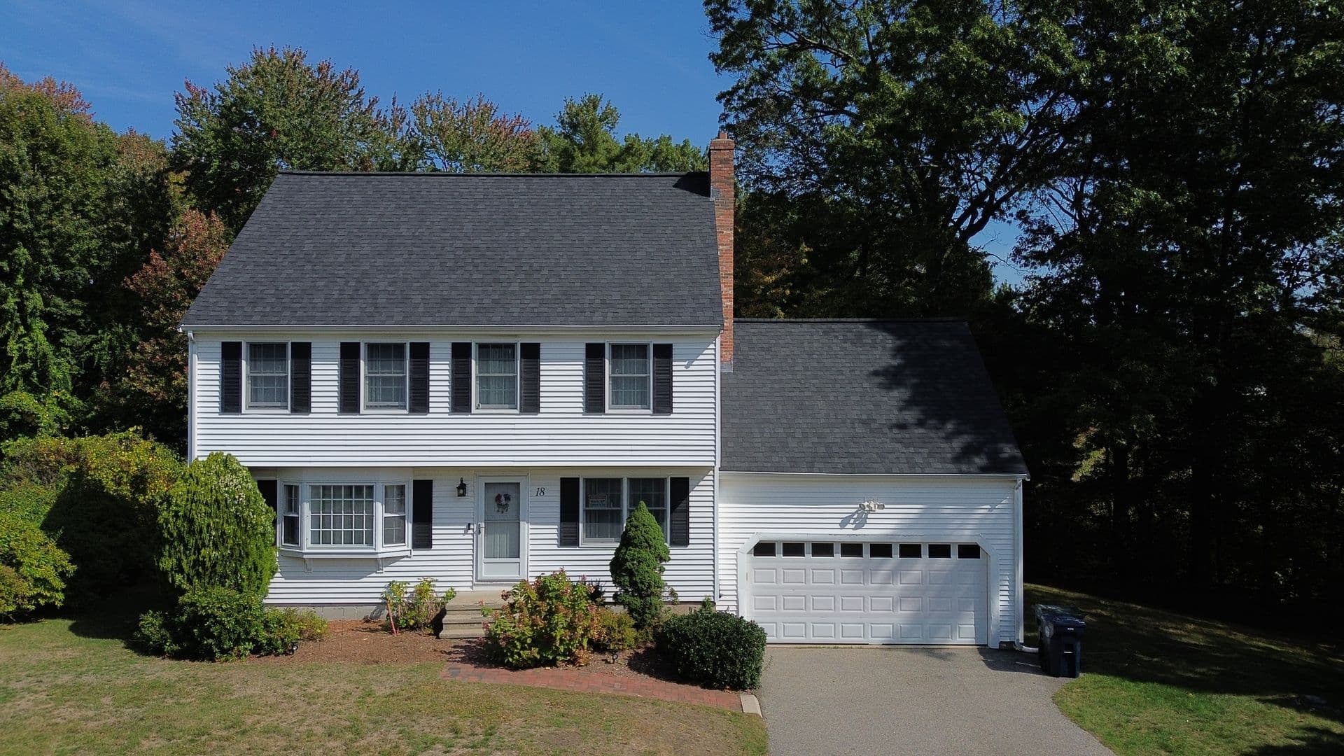 A two-story white colonial house with black shutters, a chimney, and an attached garage. It's set against a backdrop of tall trees under a clear blue sky.