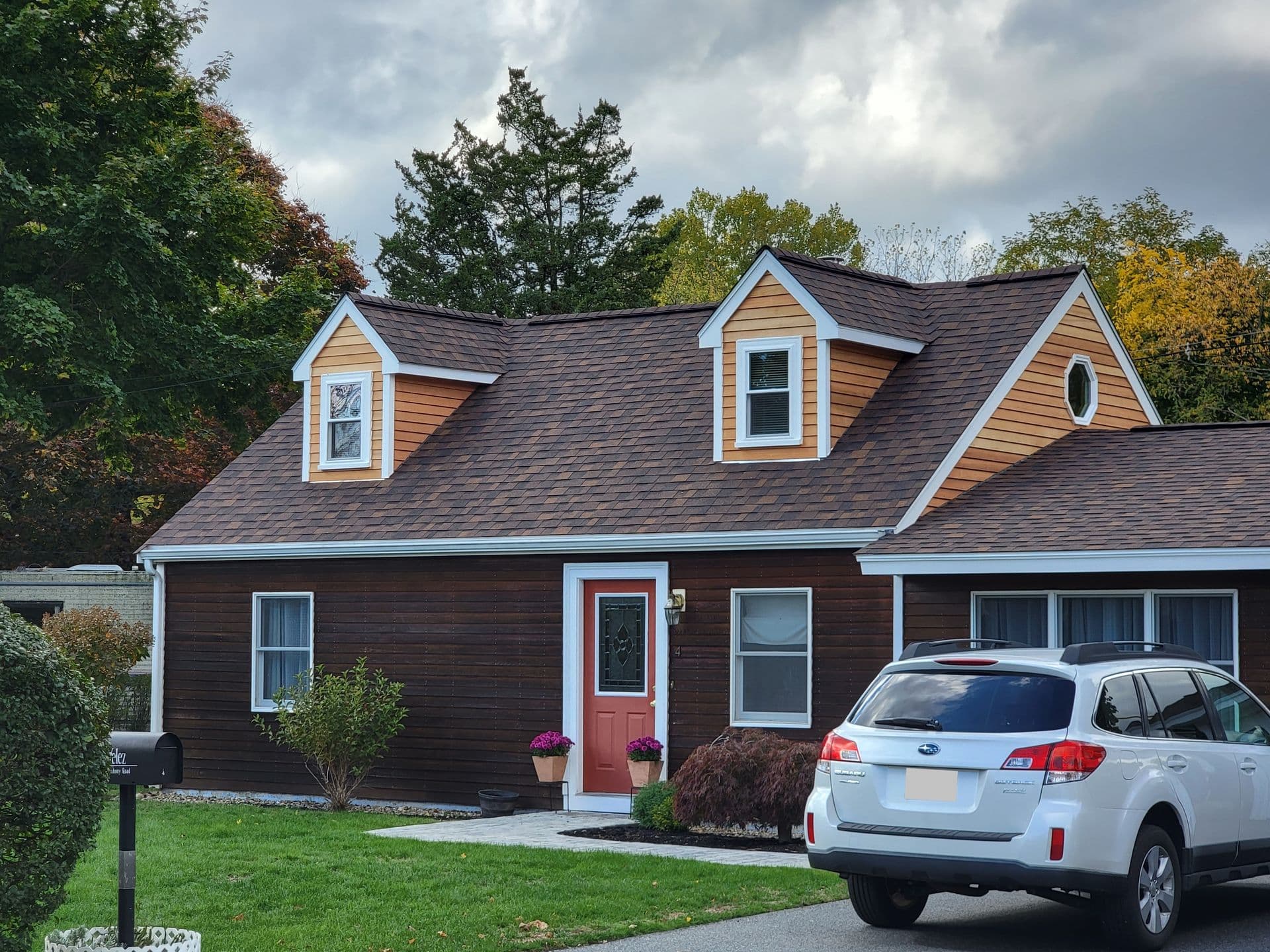 A cozy house with wooden siding and two small dormers on the roof. A white car is parked in the driveway, and potted plants adorn the entrance.