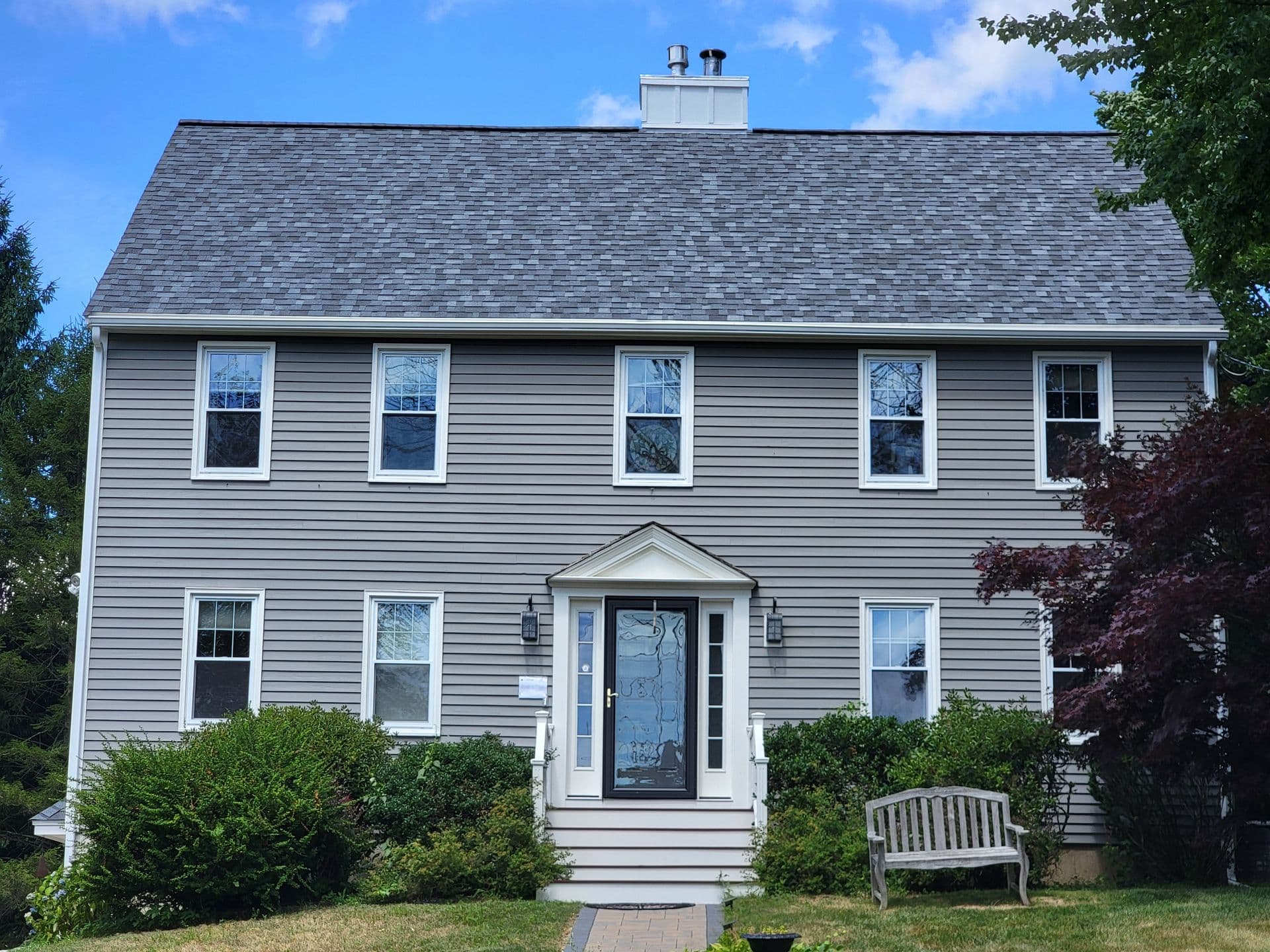 Two-story gray house with white shutters, a central door with glass, shrubs lining the entrance, and a wooden bench in front. Clear blue sky above.