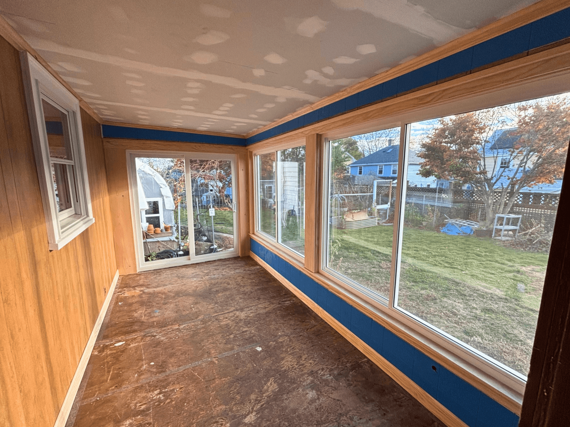 A sunroom with large windows and wood paneling overlooks a backyard. The room has an unfinished ceiling and floor, creating a work-in-progress feel.