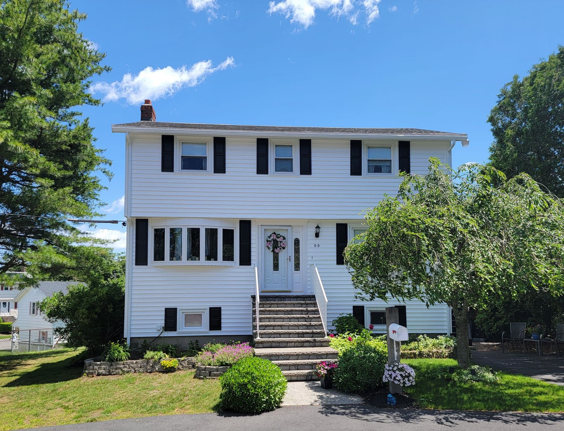 Two-story white house with black shutters, a central staircase, and a wreath on the door. Surrounded by lush greenery under a bright blue sky.
