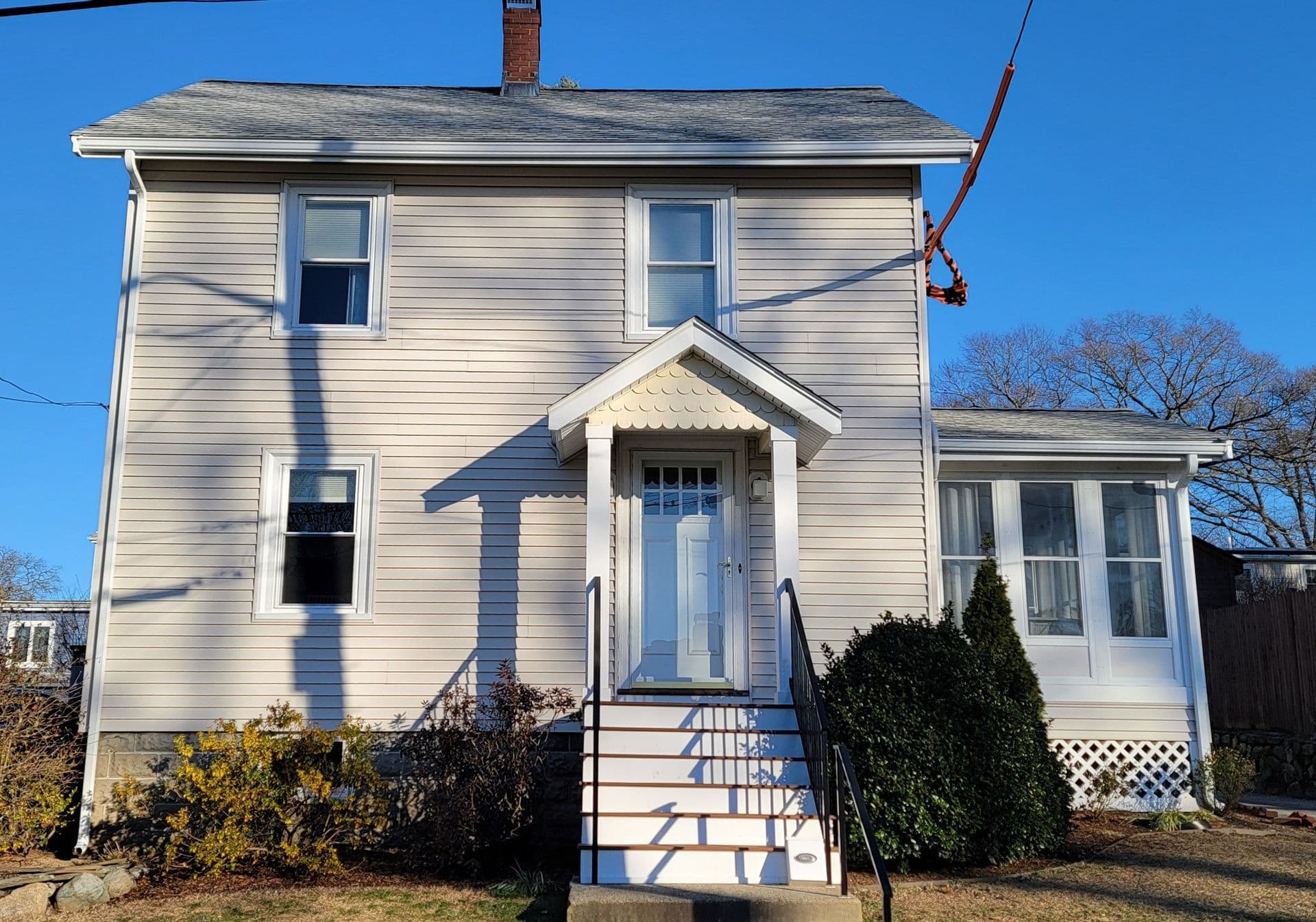 Two-story beige house with a steep roof, white trim, and a small porch. Sunlit with shadows across the front. Bushes and a lawn in the foreground.