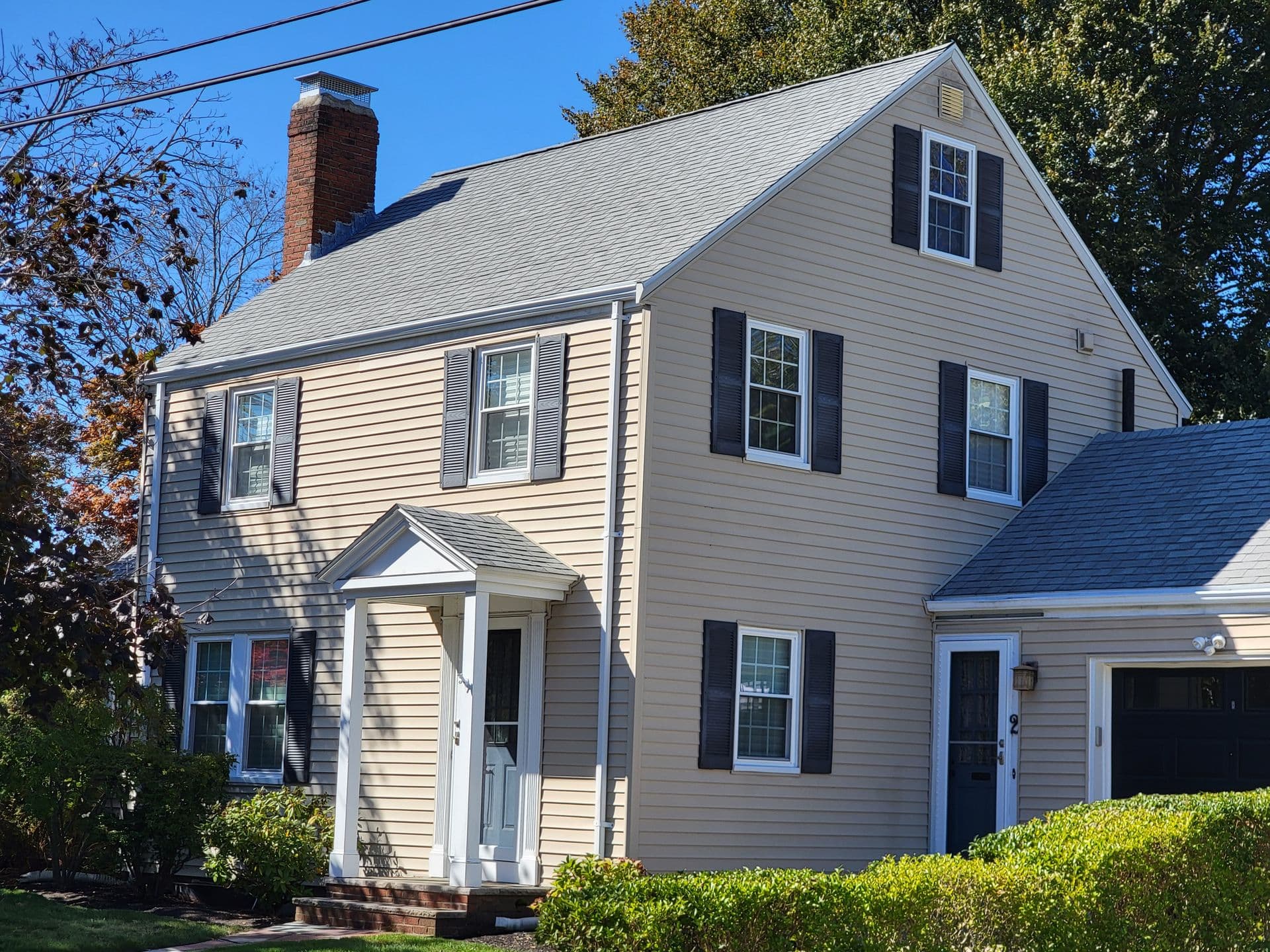 A beige two-story house with a gray roof and black shutters, featuring a white front door with a small porch. Lush greenery surrounds the home.