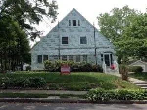 A house under renovation with exterior wrapped in weather-resistant material. A red "Realtor Sold It" sign on the front lawn. Trees and bushes surround the yard.