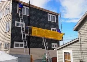 Two workers in blue jackets are on ladders renovating a gray house exterior with black wrap. A yellow tarp hangs between them against a clear sky.