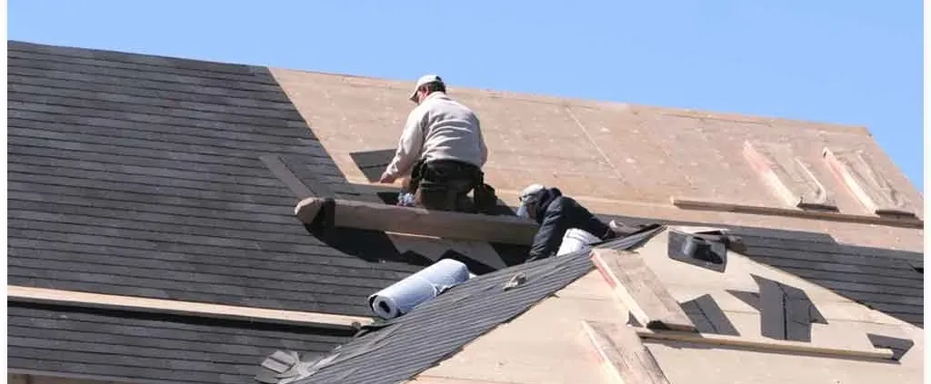 Workers installing new shingles on a roof under a clear blue sky. They are focused, wearing casual work clothing, and handling roofing materials.