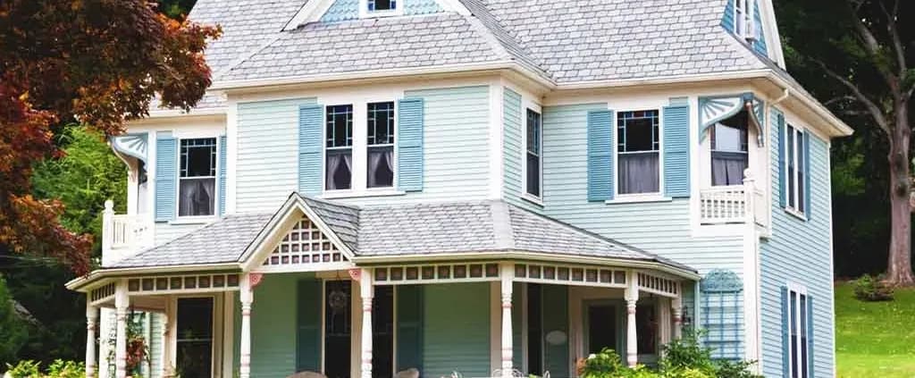 Victorian-style house with light blue clapboard siding and white trim, featuring a wraparound porch, bay windows, and lush green surroundings.