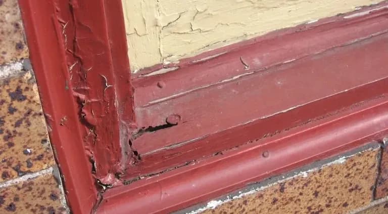 Close-up of a red wooden window frame surrounded by beige wall and brown brickwork, showing significant weathered damage and peeling paint.
