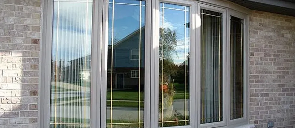 Modern bay window with reflective glass panes and white frames set in a brick wall. It reflects a blue sky and suburban houses, creating a calm atmosphere.