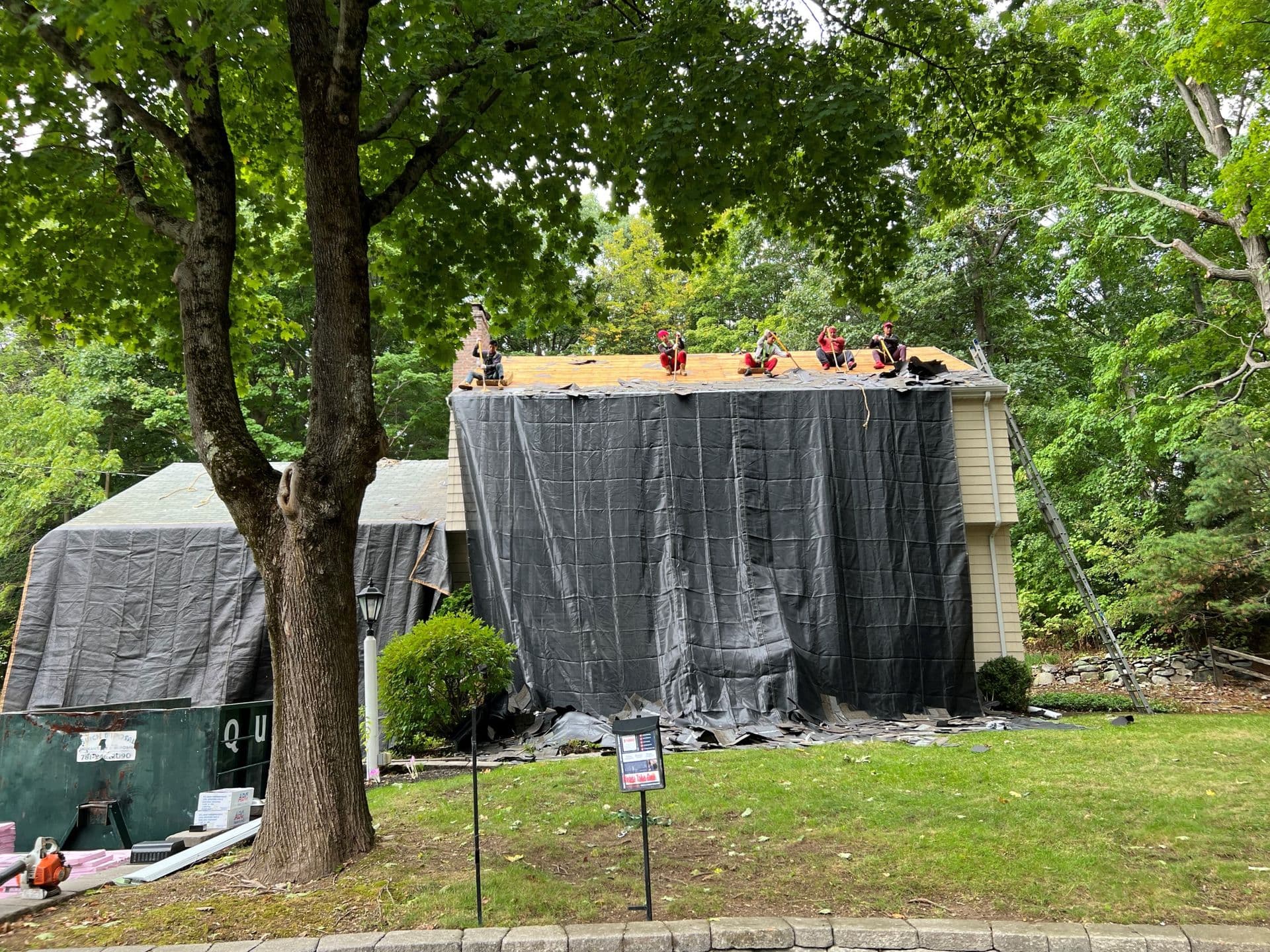 Workers on a yellow-roofed house under renovation, covered with large black tarps. Surrounded by lush trees, the scene is busy and industrious.