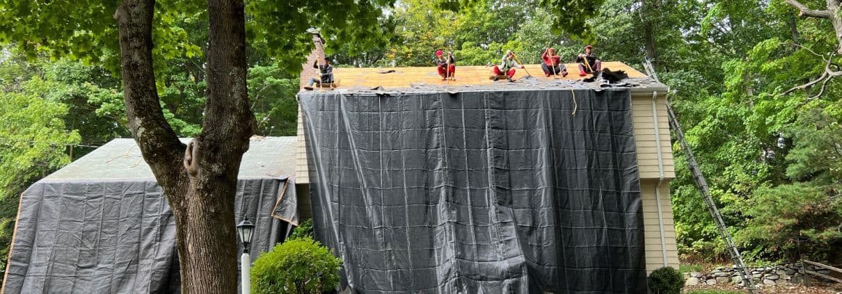 Workers on a roof install shingles, surrounded by lush trees. Large black tarps cover the house, suggesting ongoing renovation work.