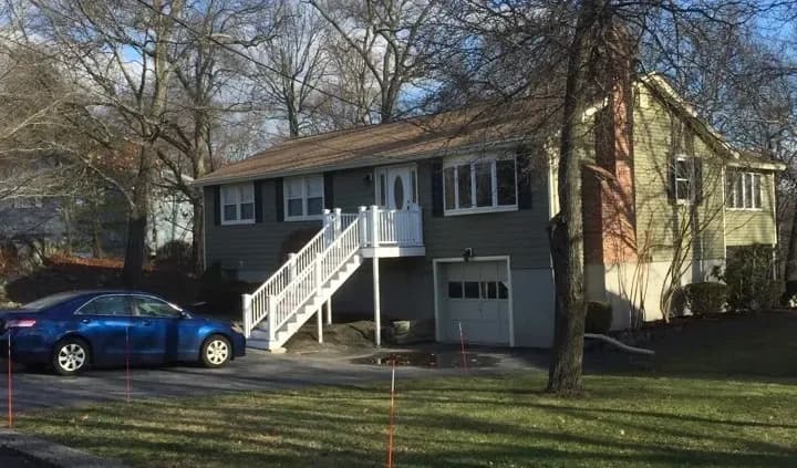 A suburban house with gray siding and a brick facade sits on a grassy lot. A set of white stairs leads to the front door. A blue car is parked in the driveway. Trees surround the property, casting bare branches against a clear sky.