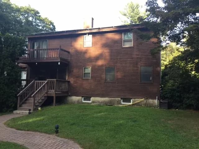 Two-story brown wooden house with a small porch and stairs, surrounded by lush greenery, under a clear sky. The setting feels calm and inviting.